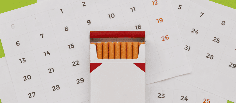 Carteira de cigarros aberta sobre folhas de calendário, representando o cálculo da carga tabágica e o histórico de consumo.