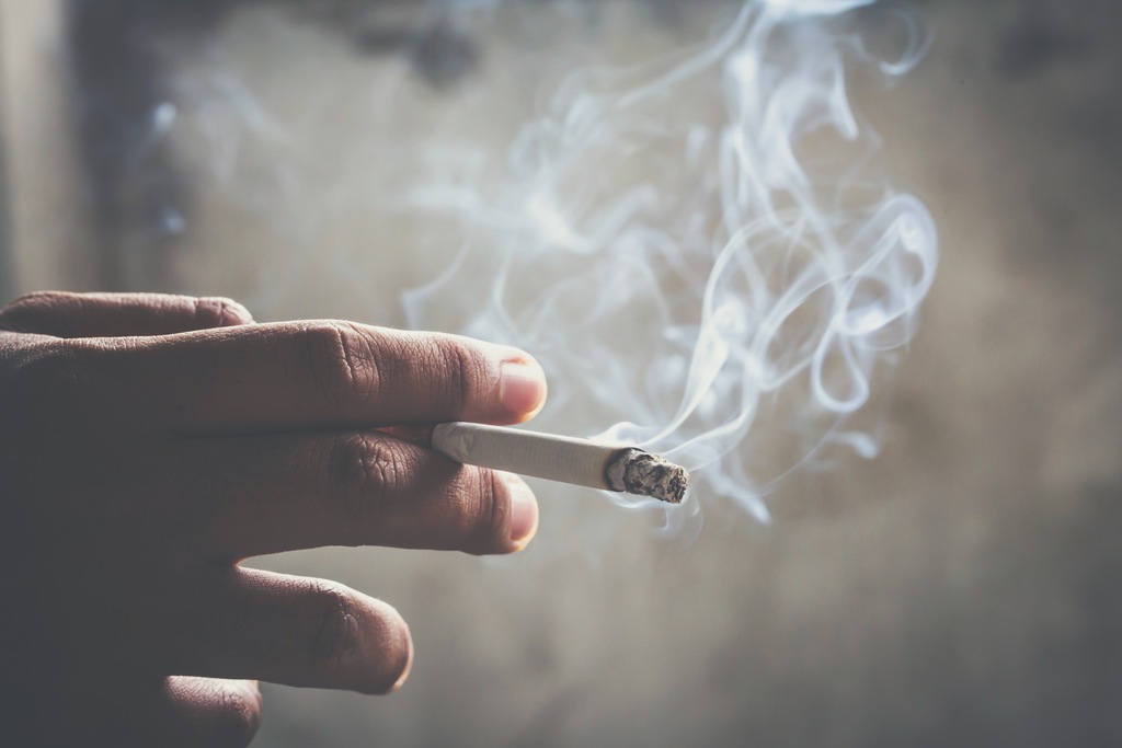 Mão segurando um cigarro aceso soltando bastante fumaça, representando os riscos do tabagismo e a relação entre câncer e cigarro.