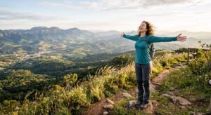 Mulher com mochila celebra com os braços abertos no topo de uma montanha com vista panorâmica. Natureza e liberdade em trilha de ecoturismo.